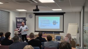 Mike O'Flynn of Samphire Marketing stands at the front of a conference room presenting to an audience. Behind him, a large screen displays a slide titled "What is the step-by-step?" with bullet points detailing steps for implementing a chatbot.