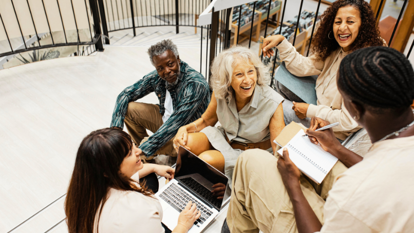 A diverse group of five adults, appearing to be of varying ages, are gathered and interacting on a staircase. One woman in the foreground with long dark hair is looking at a laptop. To her right, a person with braided hair is writing in a notebook. An older woman with white hair and another woman with curly dark hair are laughing. An older man with grey hair and a beard is also part of the group, smiling. The setting appears to be a public building with metal railings and a wide staircase.