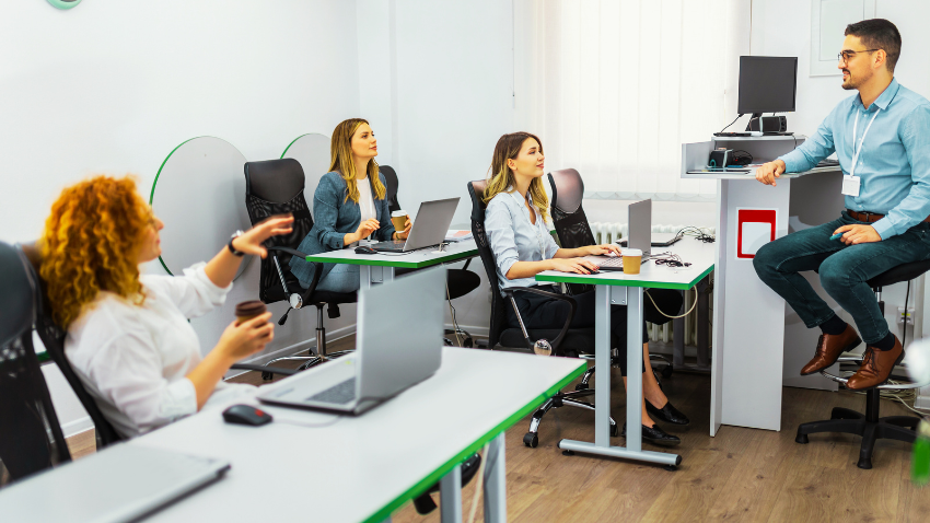 A brightly lit classroom or office space where a male instructor is speaking to three female students. The instructor, on the right, is seated on a tall stool next to a computer setup, looking towards the students. One student with long, curly red hair, on the left, is holding a coffee cup and gesturing as if asking a question. Two other students, both with long blonde hair, are seated at desks with laptops, looking towards the instructor. The desks are white with green edges.
