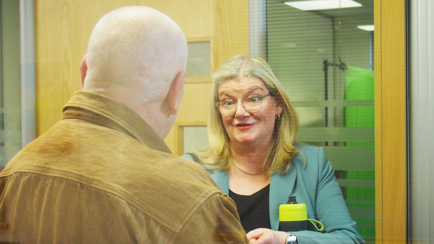 A woman with blonde hair and glasses, wearing a teal blazer and holding a yellow water bottle, is speaking to a man whose back is to the viewer. The woman is smiling and appears engaged in conversation. They are in an office setting with glass partitions and wooden door frames.