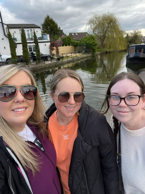 three women smiling at the camera. a selfie. background is a lake. 