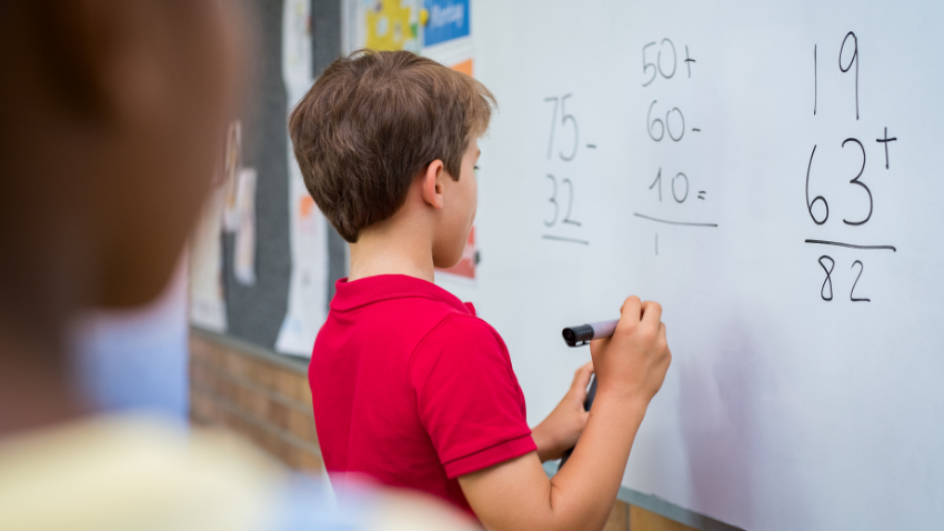 A child writing maths calculations on a whiteboard in a classroom while another pupil watches.