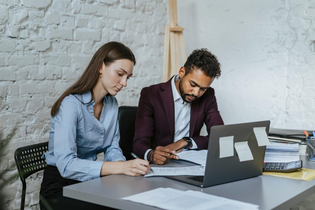 Two colleagues reviewing documents and working together at a desk with a laptop in an office setting.