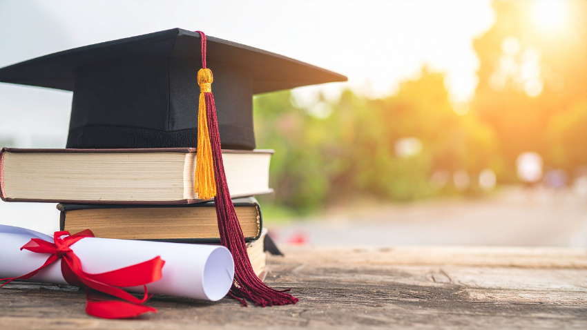 Graduation cap resting on stacked books beside a rolled diploma tied with a red ribbon, symbolising academic achievement and qualifications.